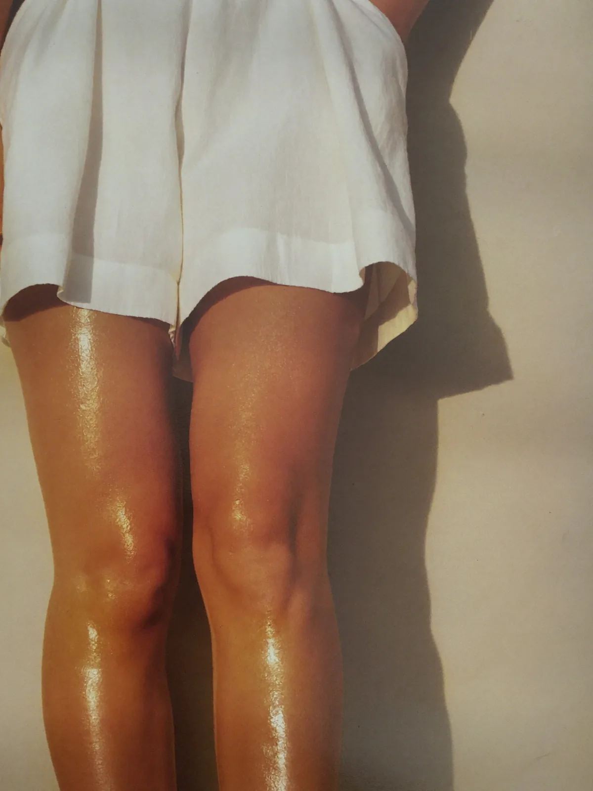 Las piernas de una mujer bajo el dobladillo de un short marfil, lisas y uniformes, con luz cálida y baja ante una pared clara